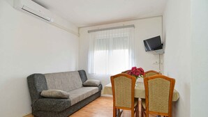 Un espace de vie compact comprenant un canapé, une table à manger, des chaises, un téléviseur fixé au mur et une fenêtre ornée de rideaux légers.