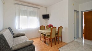 Un salon comprenant un canapé, une table à manger avec des chaises, un téléviseur à écran plat fixé au mur et une fenêtre ornée de rideaux légers.