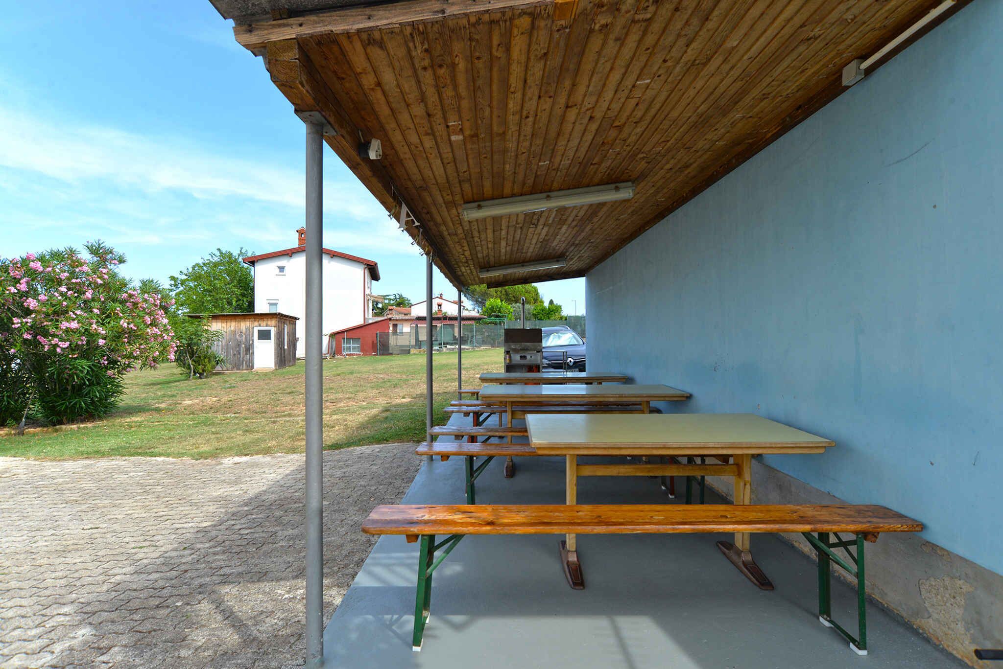Un espace extérieur aménagé avec des tables de pique-nique et des bancs, un toit abrité et une vue sur un bâtiment et la verdure en arrière-plan.