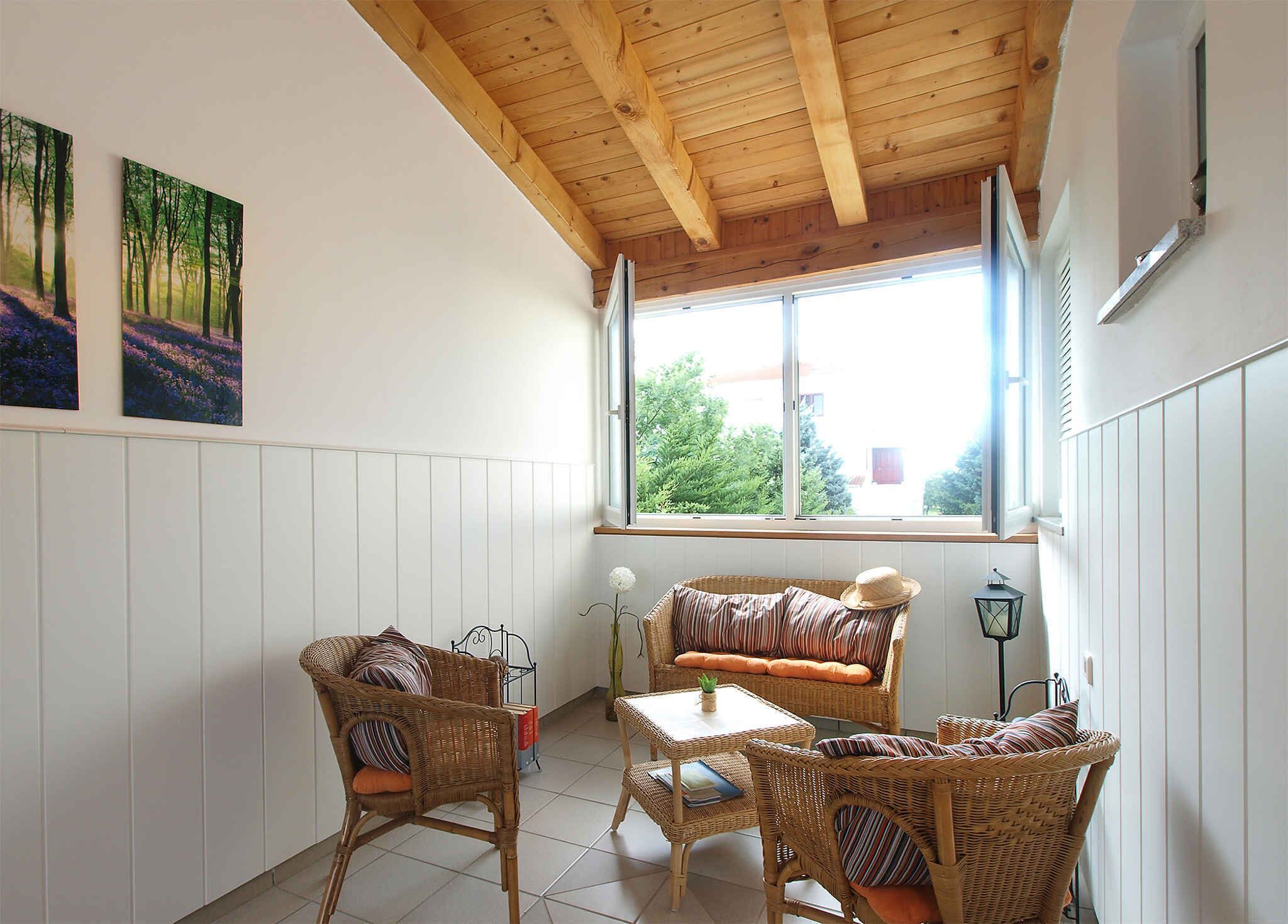 A cozy room with a wooden ceiling, a large window with a view of trees, wicker furniture, and two framed pictures on the wall.