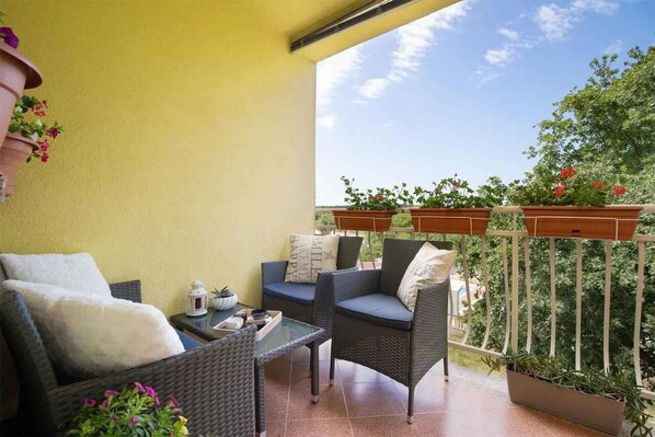 Relax on the balcony with a view of lush greenery, surrounded by a potted plant and comfortable wicker chairs.