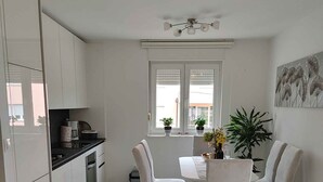 Une cuisine moderne avec des meubles blancs, une table à manger entourée de quatre chaises, une plante en pot et une fenêtre avec des stores.