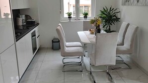 Une cuisine moderne avec des meubles blancs, une table à manger avec deux chaises et une fenêtre avec des stores.