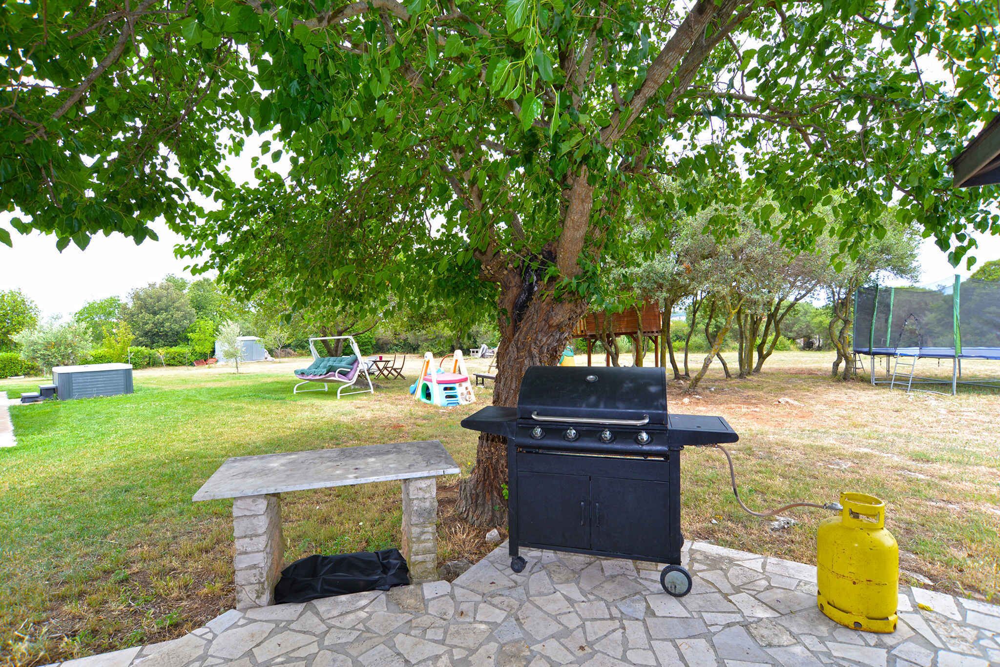 Un barbecue, une bouteille de gaz et un banc en pierre dans un espace extérieur verdoyant, agrémenté d’arbres.