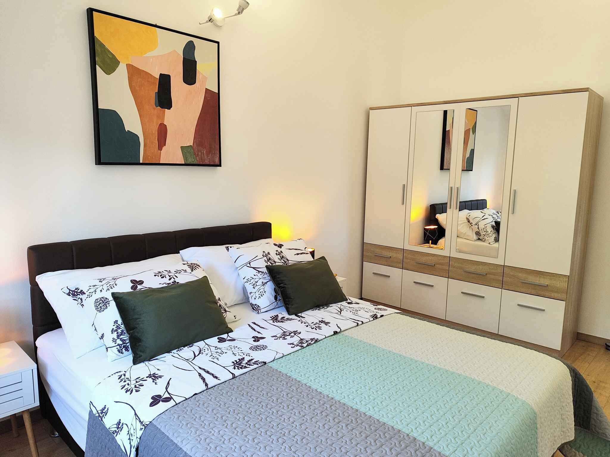 A bedroom with a bed, a wardrobe, a nightstand, and a wall-mounted abstract painting.
