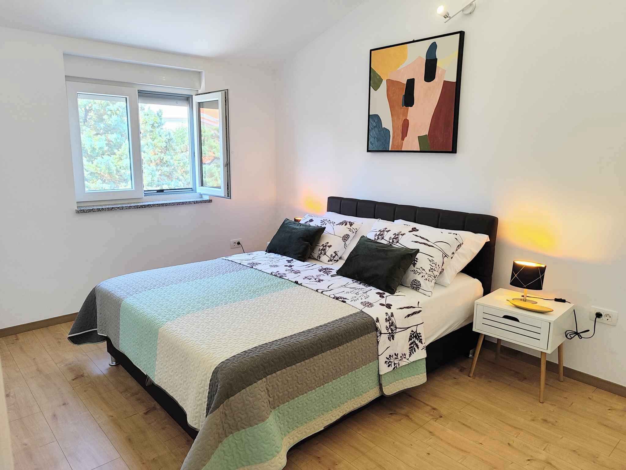 A bedroom with a bed, a nightstand, a lamp, a window, and a wall art piece.