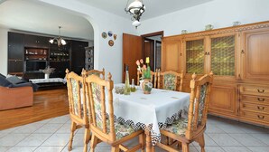 A dining area with a table set for four, surrounded by wooden chairs with floral upholstery. A wooden cabinet with glass doors is to the right, and a wooden sofa is in the background. - Holiday apartment with air conditioning (Pula)