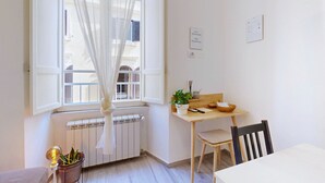 Une pièce avec une fenêtre, un radiateur, une table avec une chaise, une plante en pot et un panneau fixé au mur.