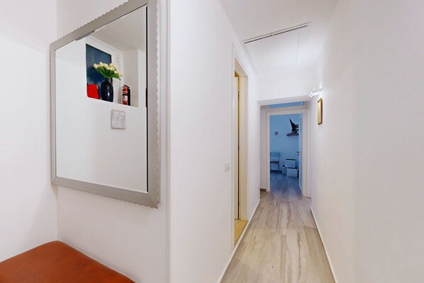 A narrow hallway with a wooden floor, a white wall with a mirror, and a door leading to another room. - Borgovi House (Roma)