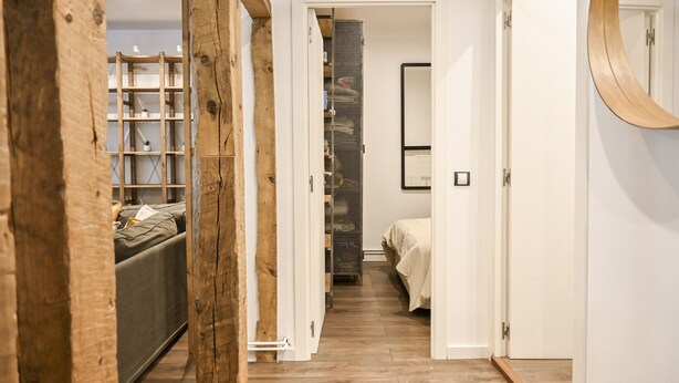 Un couloir étroit, avec une structure en bois sur le côté gauche, un placard avec des étagères, et un lit recouvert d’une couverture blanche sur le côté droit.