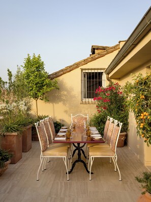 An outdoor dining area with a table set for four, surrounded by potted plants and a building with a tiled roof.