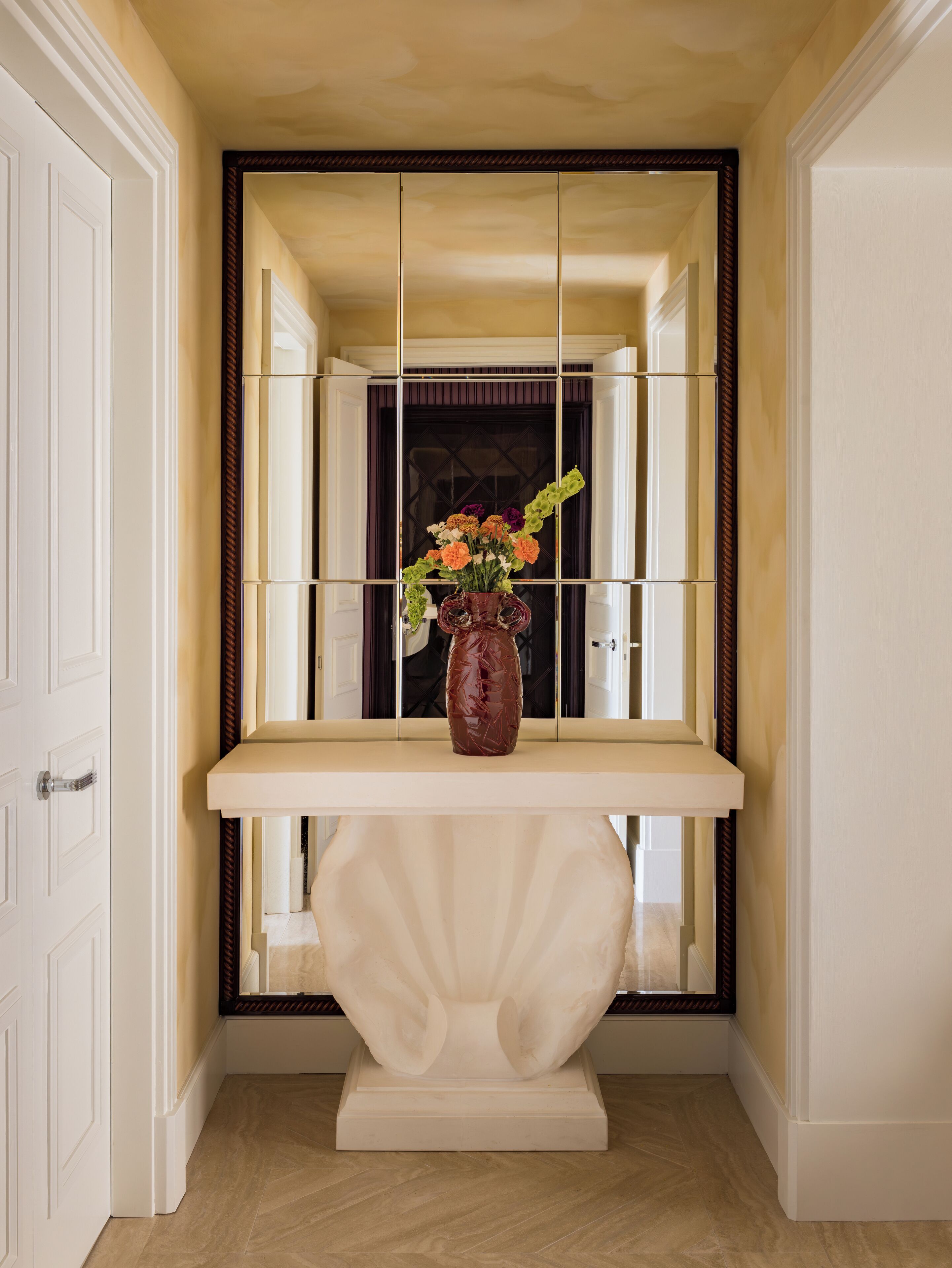 A hallway with a white door, a white console table with a vase of flowers, and a mirror on the wall.