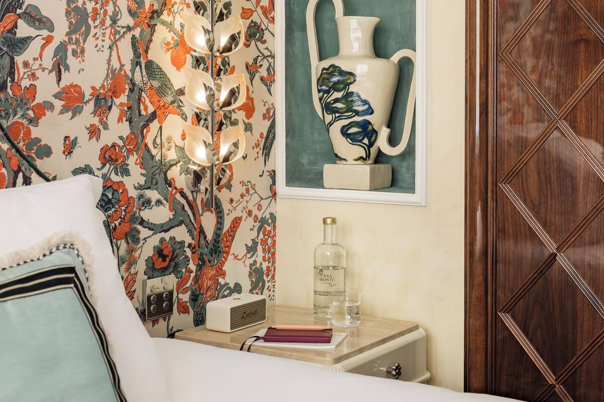 Une chambre à coucher avec un papier peint à motifs floraux, une table de chevet avec un vase, une lampe et une bouteille d’eau.