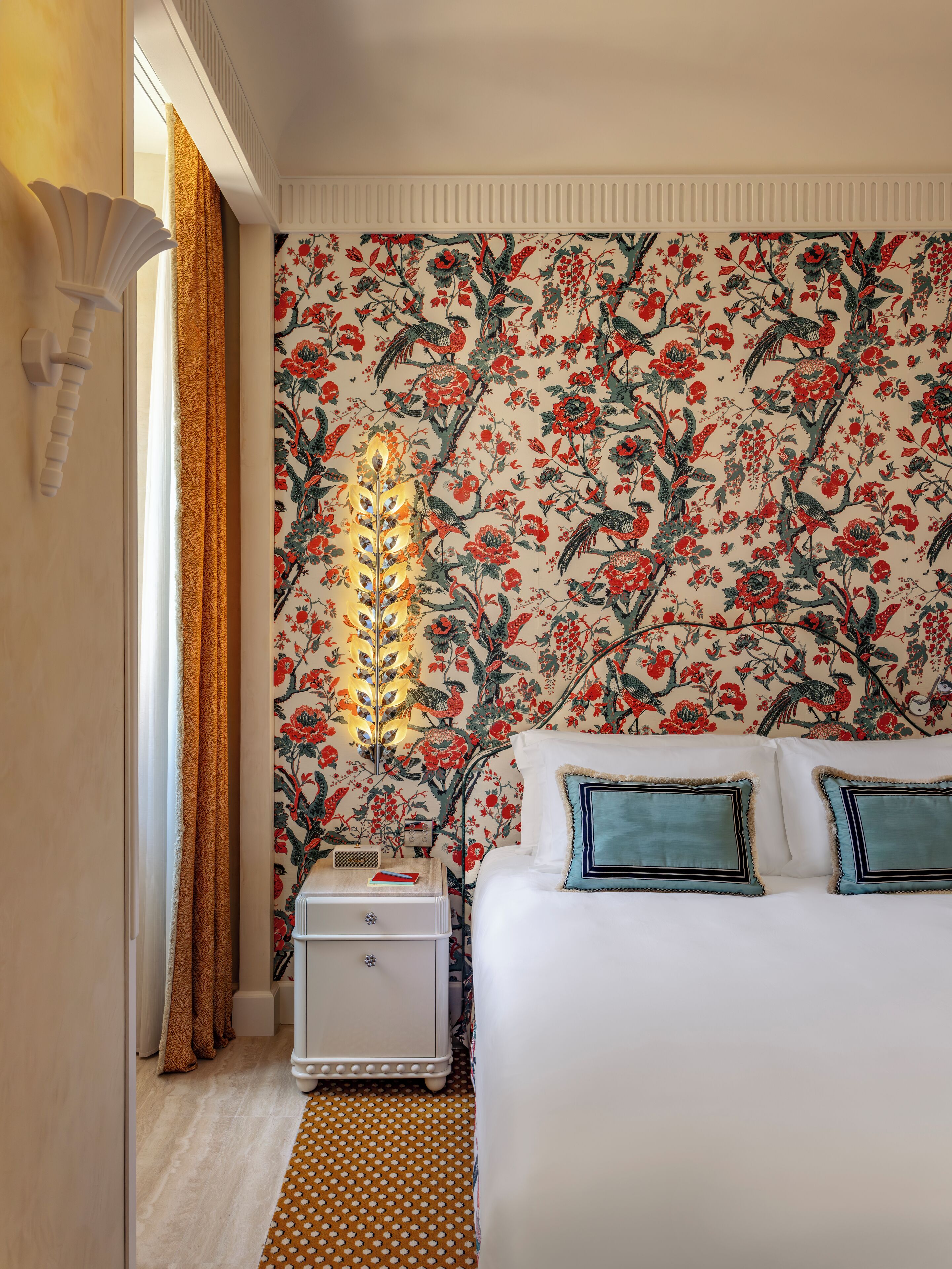 Quarto com papel de parede floral, cama branca com almofadas azul-turquesa, mesa de cabeceira com abajur e tapete estampado.