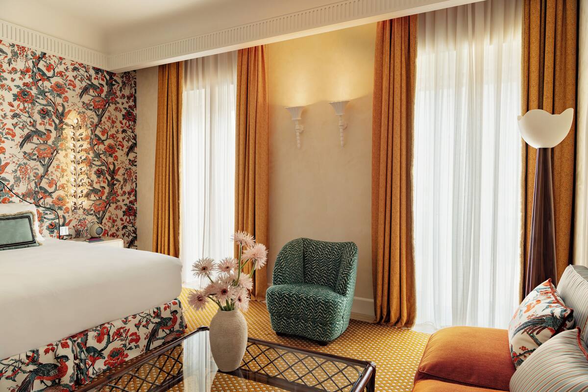 Une chambre d’hôtel avec un lit, un fauteuil vert, un canapé, un vase de fleurs et des rideaux.