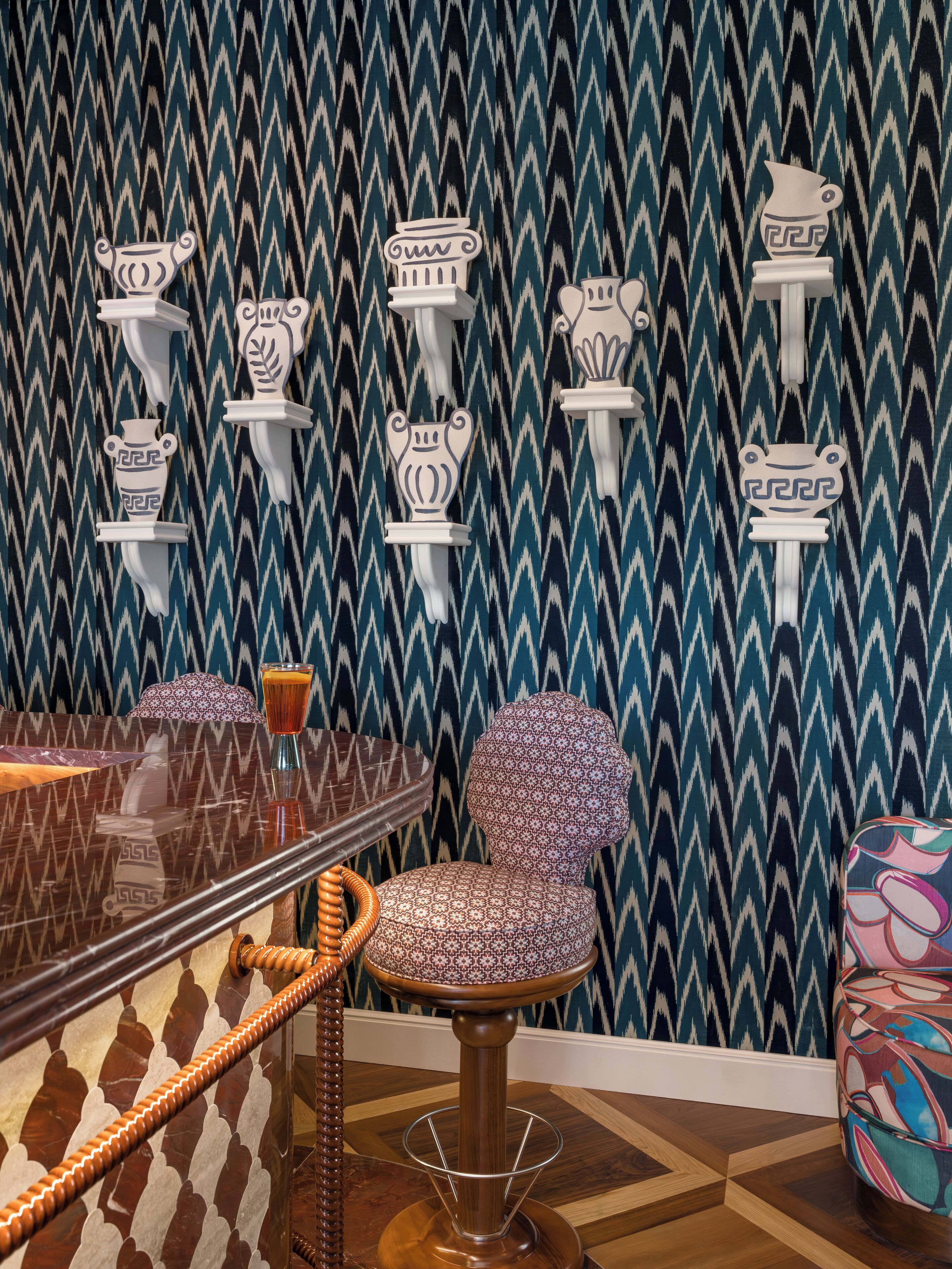 Una habitación con papel tapiz estampado, una mesa redonda con superficie de vidrio, una silla estampada y un sillón colorido.