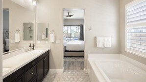 A modern bathroom with a large mirror, dual sinks, a bathtub, and a view into a bedroom. - PGA West Getaway with Pool & Fire Pit (La Quinta)