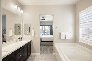 A modern bathroom with a large mirror, dual sinks, a bathtub, and a view into a bedroom. - PGA West Getaway with Pool & Fire Pit (La Quinta)