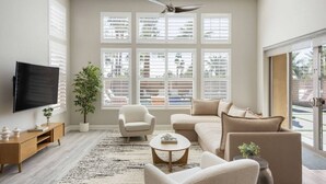 A living room with a flat-screen TV, a ceiling fan, a large window with blinds, a sofa, a coffee table, and a potted plant.