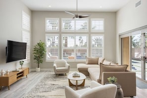 A living room with a flat-screen TV, a ceiling fan, a large window with blinds, a sofa, a coffee table, and a potted plant. - PGA West Getaway with Pool & Fire Pit (La Quinta)