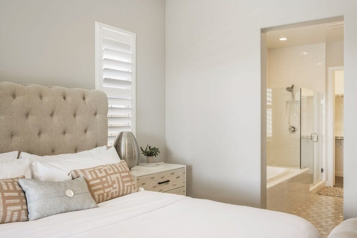A bedroom with a tufted headboard, a bedside table with a lamp, and a view into a bathroom with a shower and bathtub.