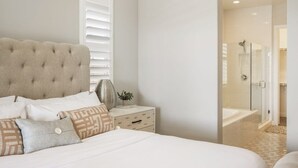 A bedroom with a tufted headboard, a bedside table with a lamp, and a view into a bathroom with a shower and bathtub. - PGA West Getaway with Pool & Fire Pit (La Quinta)