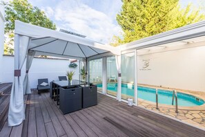 Un espace extérieur abrité comprenant une piscine, une terrasse en bois et une pièce fermée par des baies vitrées, avec un fauteuil placé au bord de la piscine.