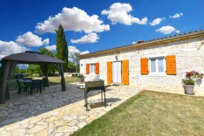 A stone building with orange shutters, a covered patio with tables and chairs, and a grassy lawn. - Holiday home with air conditioning (Debeljuhi)