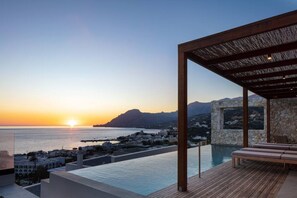 Une piscine à débordement située sur le toit, avec une terrasse en bois et des chaises longues, offrant une vue sur une ville côtière au coucher du soleil.