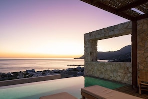 Une piscine à débordement bordée de pierres, offrant une vue sur la mer et le paysage urbain au coucher du soleil.
