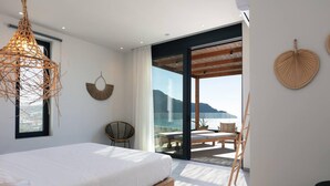 A bedroom with a bed, a wicker chandelier, a round wall hanging, and a view of the sea through a glass door. - Villa Hestia - 6 Bedroom Luxury Villa With Seaview (Agios Vasileios)