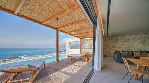 A modern interior with a wooden deck overlooking a pool and ocean, stone walls, and a dining area with a table and chairs. - Villa Hestia - 6 Bedroom Luxury Villa With Seaview (Agios Vasileios)