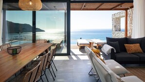 A modern living room with a large glass door leading to a balcony overlooking the sea. - Villa Hestia - 6 Bedroom Luxury Villa With Seaview (Agios Vasileios)