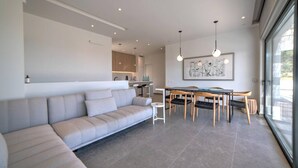 A modern living room with a sofa, dining table, and chairs. There is a kitchen area in the background. - Villa Chelidoni in Mariou (Agios Vasileios)