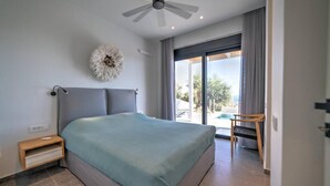 A modern bedroom with a large bed, a ceiling fan, a bedside table, and a view of the outdoors through a sliding glass door. - Villa Chelidoni in Mariou (Agios Vasileios)