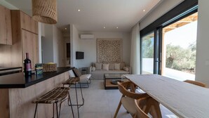 A modern kitchen with wooden cabinetry, a bar area, and a dining space with a large window. - Villa Mare in Mariou (Agios Vasileios)