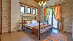 A bedroom with a stone wall, a wooden ceiling, a metal bed frame, and a view through a window with curtains. - Villa Helios in Foinikas (Agios Vasileios)