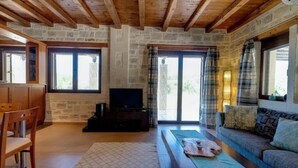 A cozy living room with a stone wall, wooden beams, a TV, a sofa, and a dining area. - Villa Helios in Foinikas (Agios Vasileios)