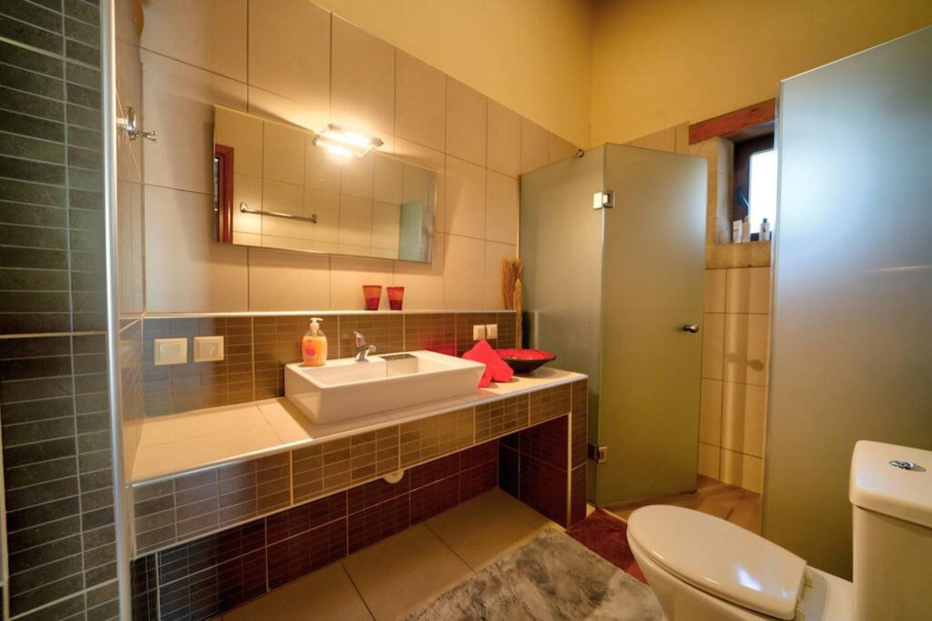 Een badkamer met een wit wastafel, spiegel en toilet. Er is een handdoekhouder boven het wastafel en een douchegedeelte aan de rechterkant.