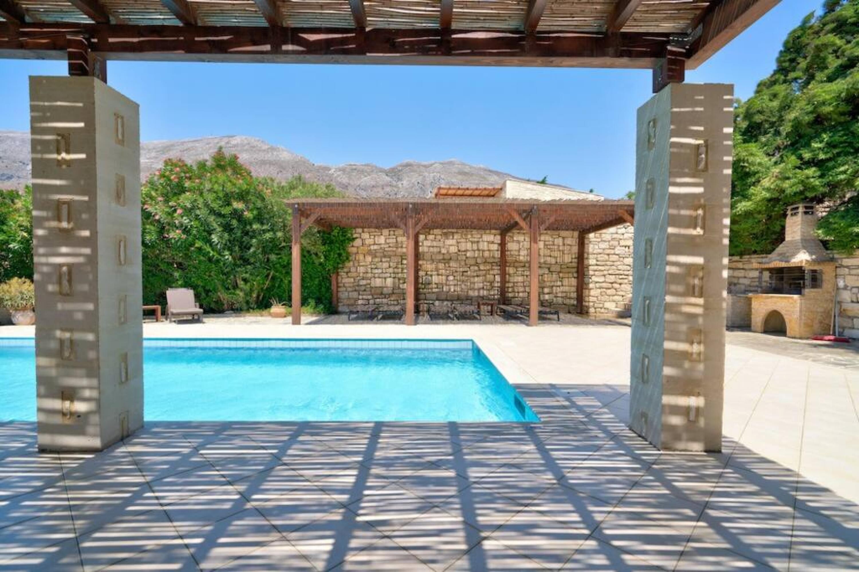 A swimming pool with a pergola structure, stone walls, and a view of mountains in the background.