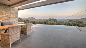 A modern outdoor patio with a stone wall, wicker furniture, and a view of a mountain landscape. - Villa Oniriko in Mariou (Agios Vasileios)