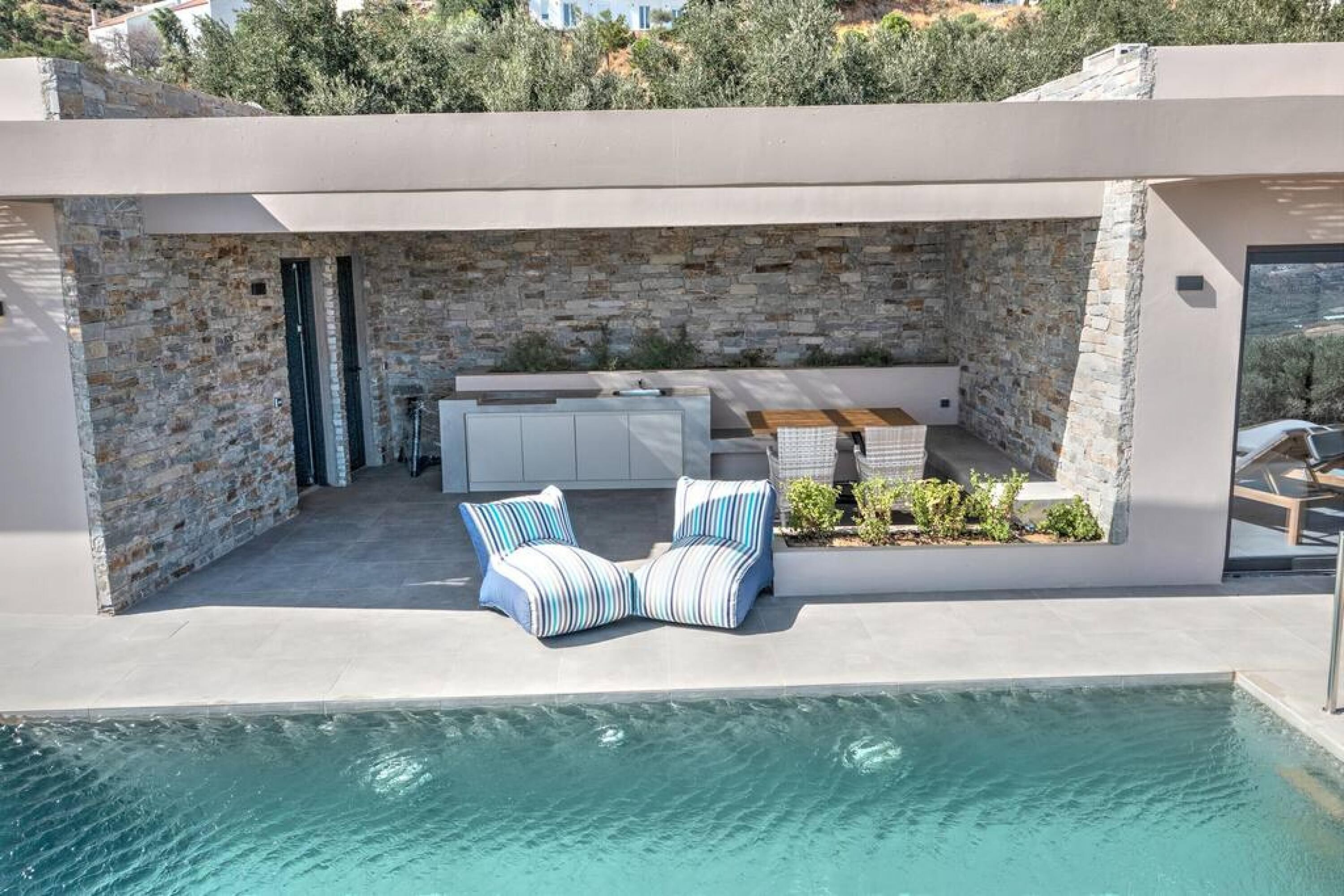 A modern outdoor pool area with stone walls, a seating area with striped cushions, and a kitchen with white cabinets.