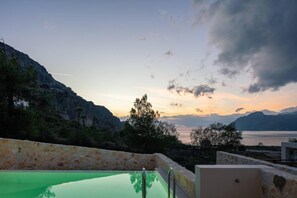 A swimming pool with a stone wall, overlooking a mountainous landscape and a body of water during sunset.