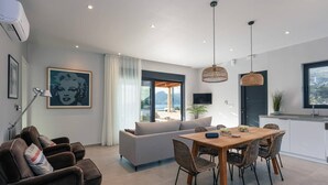 A modern living room with a sofa, dining table, and chairs. There is a large window with a view of the outdoors. - Villa Petra in Plakias (Agios Vasileios)
