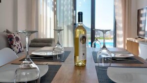 A modern dining area with a wooden table set for two, a bottle of white wine, and a glass. The room has large windows with curtains, a sofa, and a TV. - Villa Iris in Mariou (Agios Vasileios)