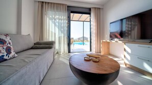 A modern living room with a sofa, a coffee table, a TV, and a view of a swimming pool. - Villa Iris in Mariou (Agios Vasileios)