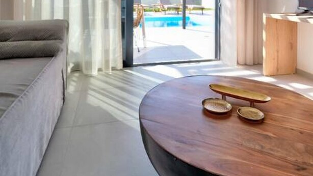 A modern hotel room with a large sliding glass door leading to a pool area, a round wooden coffee table, and a sofa. - Villa Iris in Mariou (Agios Vasileios)