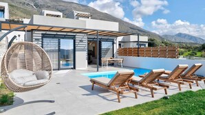 A modern outdoor pool area with lounge chairs, a hanging chair, and a glass-enclosed patio. - Villa Iris in Mariou (Agios Vasileios)