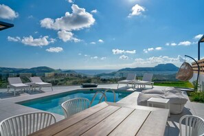 Ein Poolbereich mit Liegestühlen und einem Tisch, umgeben von einer malerischen Aussicht auf Berge und Meer.