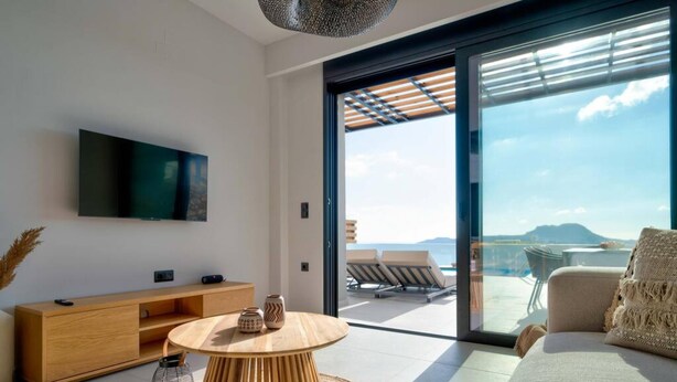 A modern living room with a round wooden coffee table, a flat-screen TV mounted on the wall, and a large sliding glass door leading to a balcony with a view of the sea and mountains. - Villa Saturday in Foinikas (Agios Vasileios)